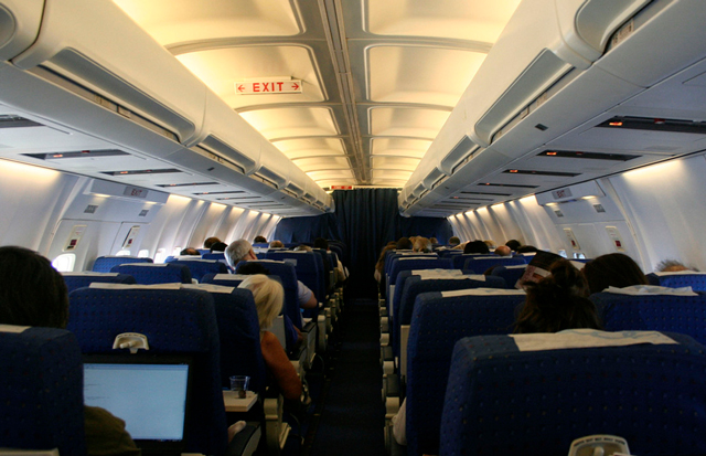 Imagem do interior de um avião assentos e passageiros sentados e luzes acesas.