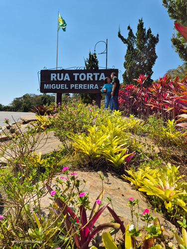 Rua torta com muitas plantas e uma placa escrita "Rua Torta Miguel Pereira"
